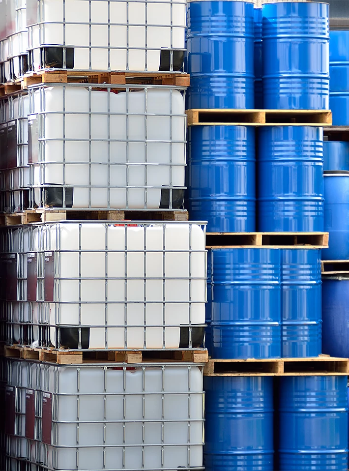 Plastic tank containers and metal drums of chemicals stacked in warehouse. 